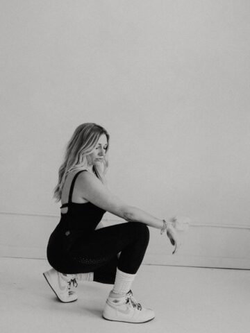 Black-and-white photo of a female sports dietitian squatting in a minimalist studio, wearing a black athletic set and white sneakers, representing strength, body recomposition, and muscle-building after three kids.