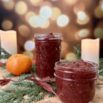 Two glass jars of homemade cranberry chia jam sitting on a rustic wooden board, surrounded by cedar branches, a clementine, and soft glowing candles — a cozy, holiday-inspired setup that highlights the rich red color and texture of the jam.
