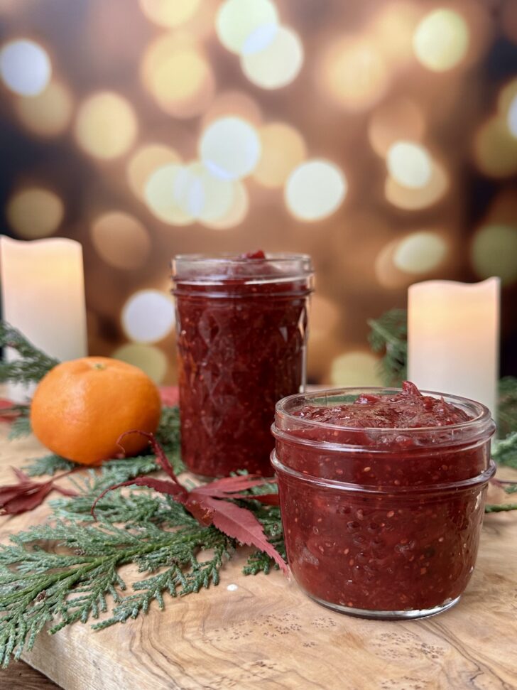 Two glass jars of homemade cranberry chia jam sitting on a rustic wooden board, surrounded by cedar branches, a clementine, and soft glowing candles. A cozy, holiday-inspired setup that highlights the rich red color and texture of the jam.