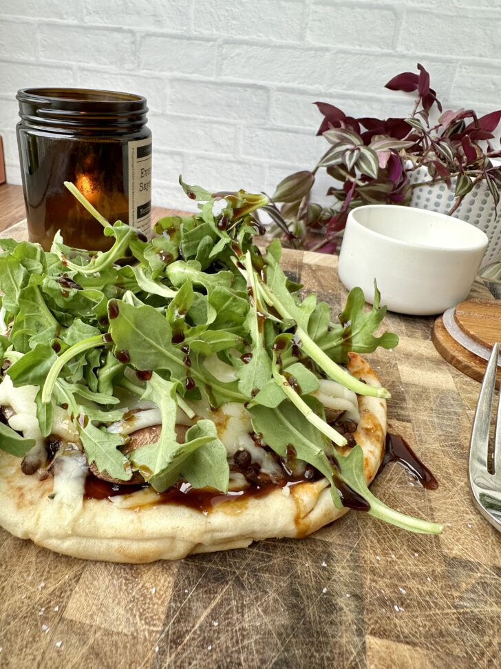 High-protein pita pizza topped with arugula, melted cheese, lentils, and balsamic glaze on a wooden cutting board