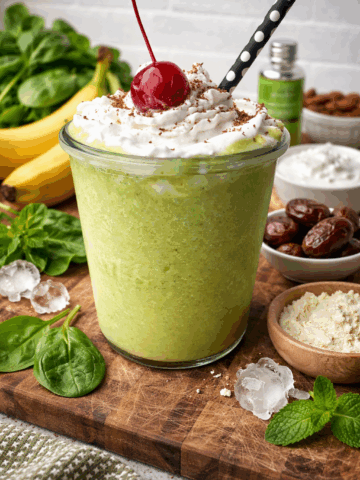 High-protein Shamrock shake in a clear glass jar topped with coconut whipped cream, chocolate shavings, and a maraschino cherry, surrounded by fresh spinach, banana, dates, vanilla protein powder, peppermint extract, and ice on a rustic wooden board against a light brick background.