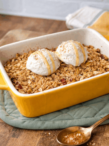 High-protein banana foster cobbler baked in a yellow ceramic dish topped with two scoops of Greek yogurt and maple drizzle on a wooden table.