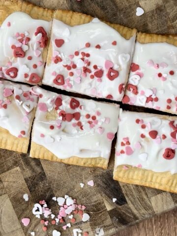 Giant homemade raspberry pop tart topped with vanilla Greek yogurt frosting and Valentine’s Day heart sprinkles, sliced into squares on a wooden board.