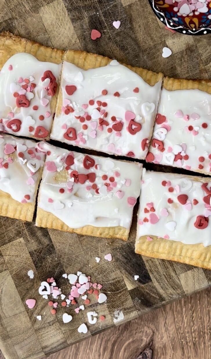 Giant homemade raspberry pop tart topped with vanilla Greek yogurt frosting and Valentine’s Day heart sprinkles, sliced into squares on a wooden board.