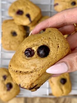 High-protein lemon blueberry breakfast cake held in hand with fresh blueberries baked on top, cooling on a wire rack in the background.