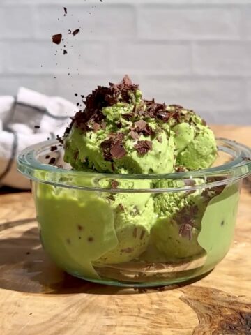 Creamy high-protein mint chocolate chip ice cream made with blended cottage cheese and dark leafy greens, topped with shaved dark chocolate in a clear glass bowl.
