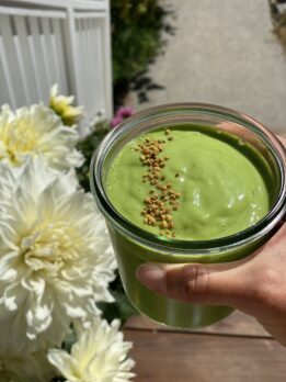 Creamy tropical green smoothie made with kale, spinach, mango, and coconut water, topped with bee pollen in a glass jar outdoors in natural sunlight.