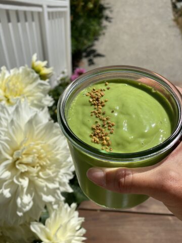 Creamy tropical green smoothie made with kale, spinach, mango, and coconut water, topped with bee pollen in a glass jar outdoors in natural sunlight.