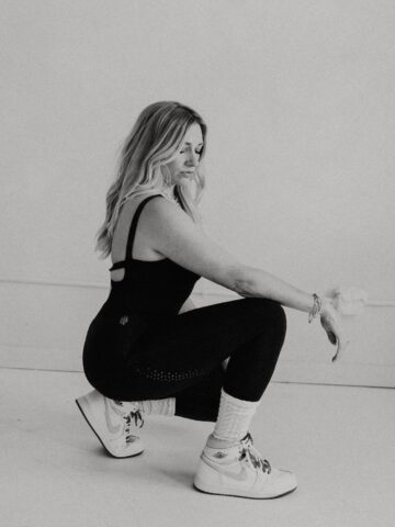 Black-and-white photo of a female sports dietitian squatting in a minimalist studio, wearing a black athletic set and white sneakers, representing strength, body recomposition, and muscle-building after three kids.