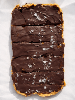 High-protein no-bake peanut butter bars topped with a thick layer of dark chocolate and flaky sea salt, sliced into rectangles and photographed on parchment paper in natural light.