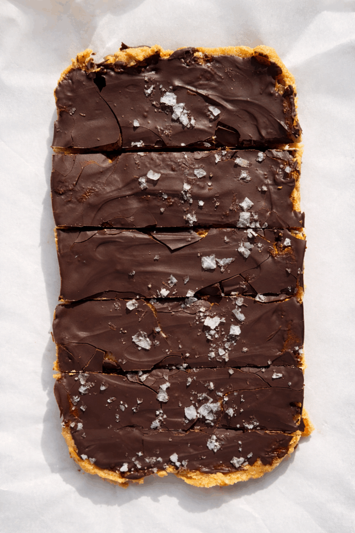 High-protein no-bake peanut butter bars topped with a thick layer of dark chocolate and flaky sea salt, sliced into rectangles and photographed on parchment paper in natural light.