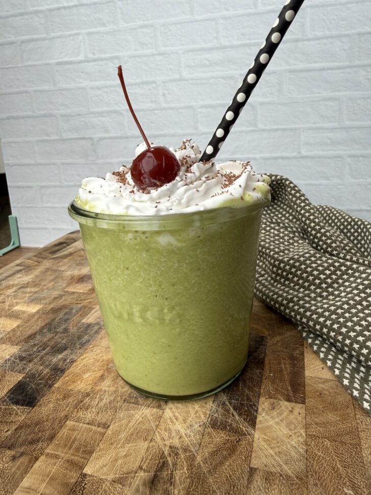 High-protein Shamrock Shake in a clear glass jar topped with coconut whipped cream, chocolate shavings, and a maraschino cherry, with a black polka dot straw, set on a wooden cutting board against a light brick background with a green patterned cloth.