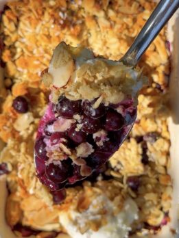Close-up of a spoonful of high-protein blueberry orange crumble with juicy baked blueberries, golden oat topping, sliced almonds, and creamy Greek yogurt in a white baking dish.