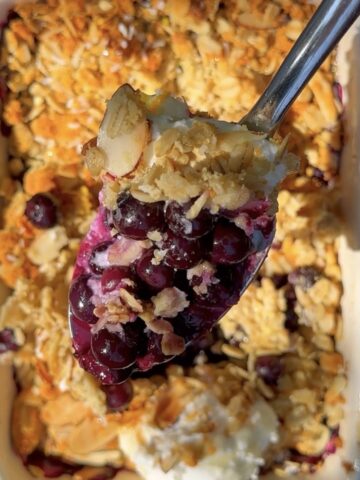 Close-up of a spoonful of high-protein blueberry orange crumble with juicy baked blueberries, golden oat topping, sliced almonds, and creamy Greek yogurt in a white baking dish.