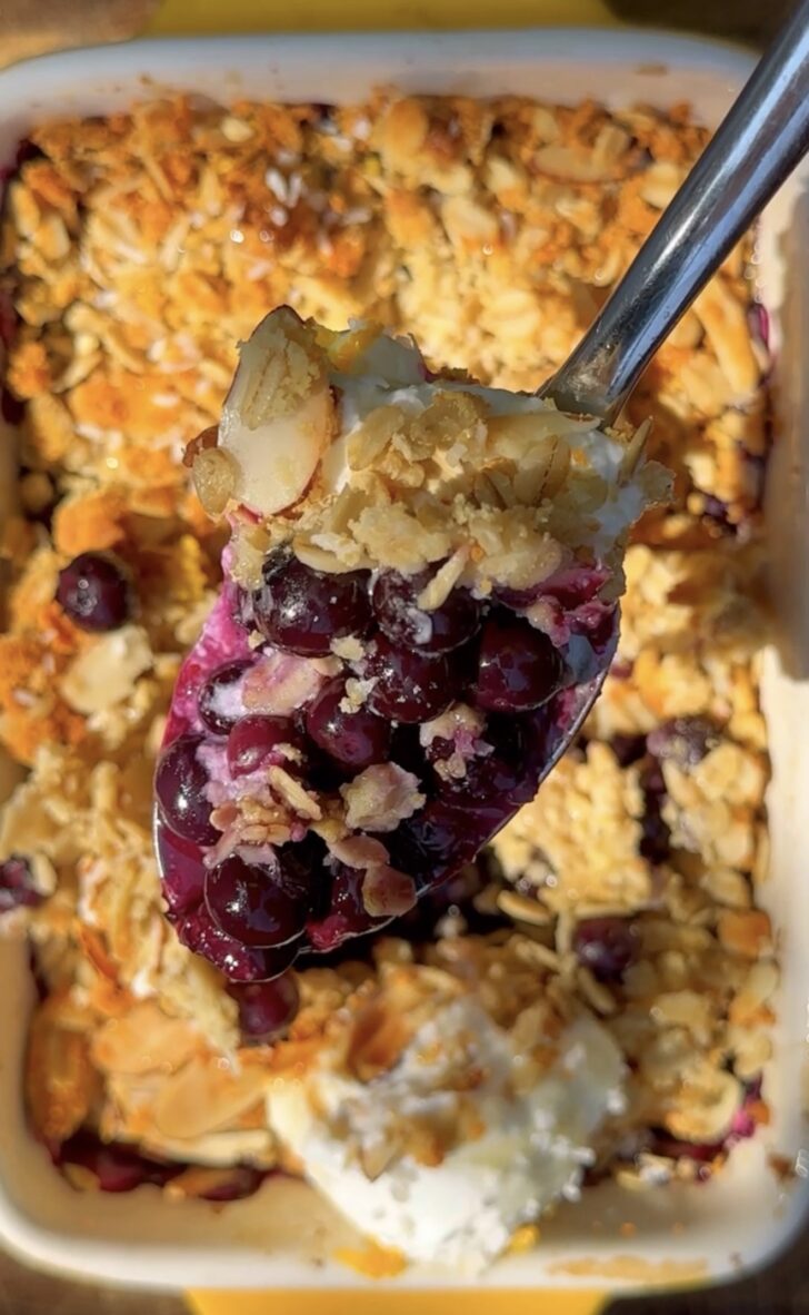 Close-up of a spoonful of high-protein blueberry orange crumble with juicy baked blueberries, golden oat topping, sliced almonds, and creamy Greek yogurt in a white baking dish.