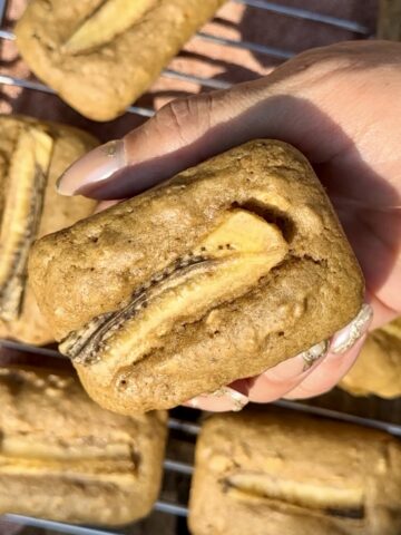 Close-up of a hand holding a soft, golden-brown banana bread breakfast cake with a baked banana slice on top, resting on a cooling rack with more mini cakes in the background.