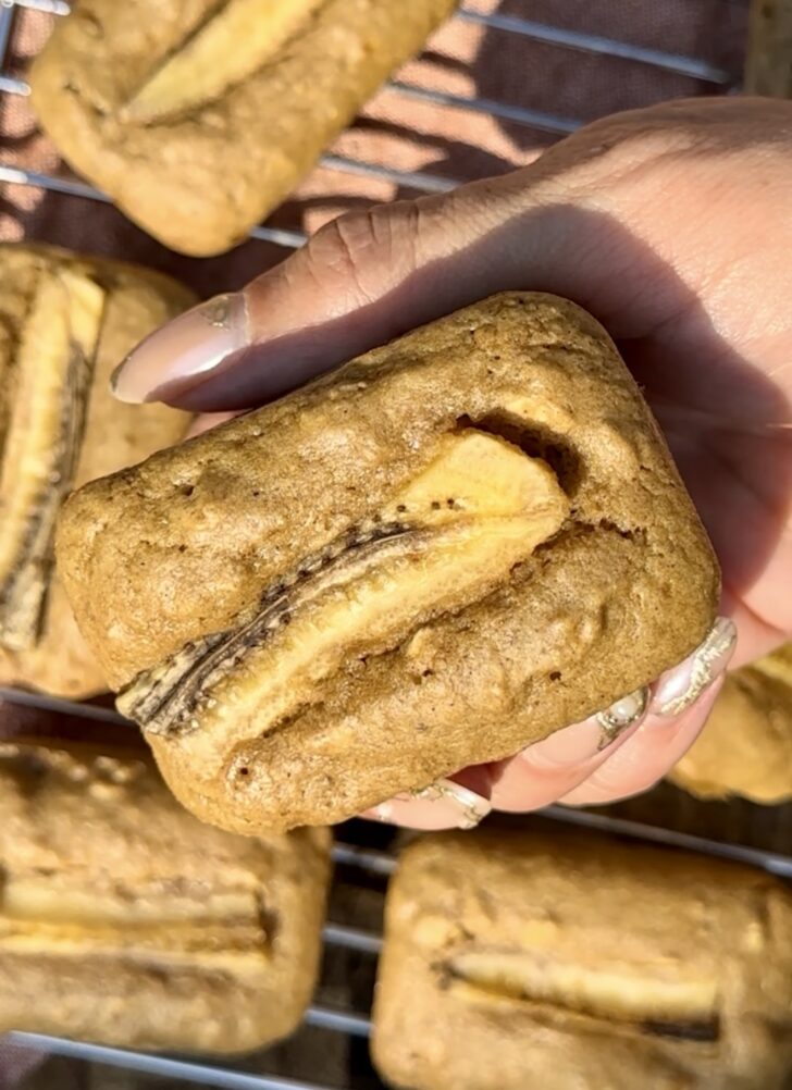 Close-up of a hand holding a soft, golden-brown banana bread breakfast cake with a baked banana slice on top, resting on a cooling rack with more mini cakes in the background.