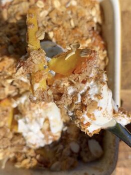 High-protein peach cobbler with oats and Greek yogurt, close-up of a spoonful showing juicy baked peaches, crumb topping, and creamy yogurt drizzle