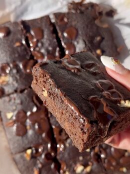 Close-up of a thick, fudgy chocolate brownie square with melted chocolate chips and a glossy drizzle on top, being held by hand, with more cut brownies scattered in the background.