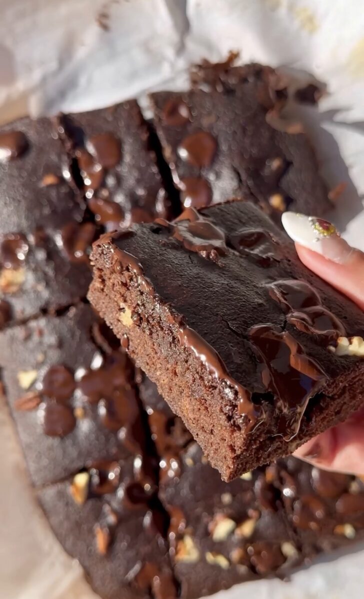 Close-up of a thick, fudgy chocolate brownie square with melted chocolate chips and a glossy drizzle on top, being held by hand, with more cut brownies scattered in the background.