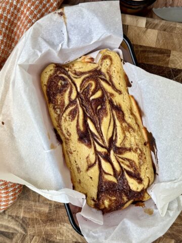 Overhead view of a freshly baked cinnamon swirl cottage cheese bread in a parchment-lined loaf pan, featuring a golden surface with dark cinnamon swirls, slightly caramelized edges, and a soft, moist texture, set on a wooden cutting board with a cozy kitchen towel nearby.