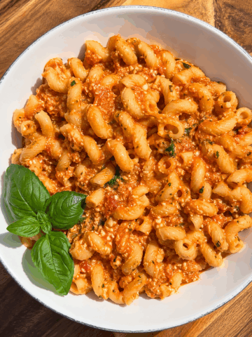 Creamy baked pink pasta made with high-protein elbow pasta tossed in a rich roasted tomato and cottage cheese sauce, garnished with fresh basil in a white bowl on a rustic wooden table. Cozy healthy comfort food recipe perfect for easy weeknight dinners, meal prep, and high-protein meals.