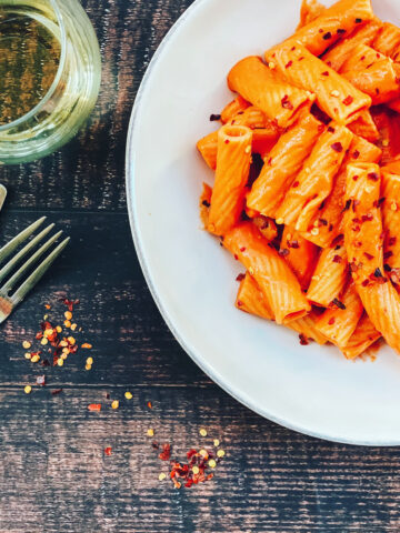 Creamy plant-based rigatoni vodka pasta served on a white plate with crushed red pepper flakes, styled on a dark rustic table with a fork and a small bowl of seasoning nearby.