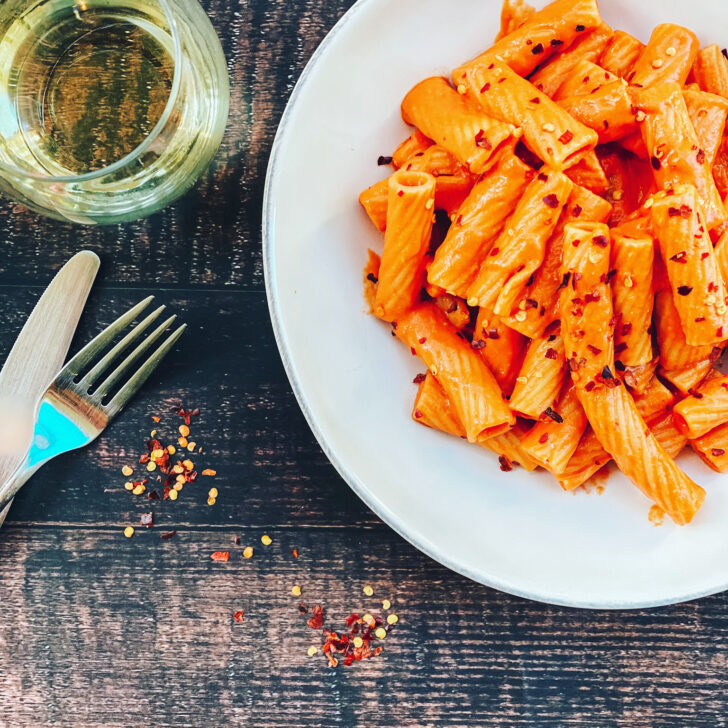 Creamy plant-based rigatoni vodka pasta served on a white plate with crushed red pepper flakes, styled on a dark rustic table with a fork and a small bowl of seasoning nearby.