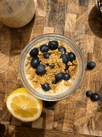 Overhead photo of lemon blueberry cheesecake overnight oats in a glass jar on a rustic wooden cutting board. The oats are topped with fresh blueberries, crushed graham crackers, and a creamy cheesecake layer, with half a lemon placed beside the jar in warm natural sunlight.