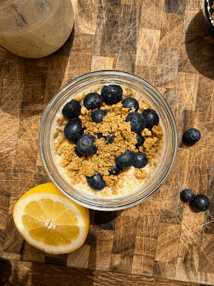 Overhead photo of lemon blueberry cheesecake overnight oats in a glass jar on a rustic wooden cutting board. The oats are topped with fresh blueberries, crushed graham crackers, and a creamy cheesecake layer, with half a lemon placed beside the jar in warm natural sunlight.