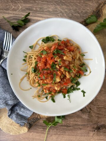 Overhead photo of maple tempeh marinara pasta served in a white bowl with chickpea spaghetti, marinara sauce, crumbled tempeh, fresh parsley, and herbs on a wooden table.