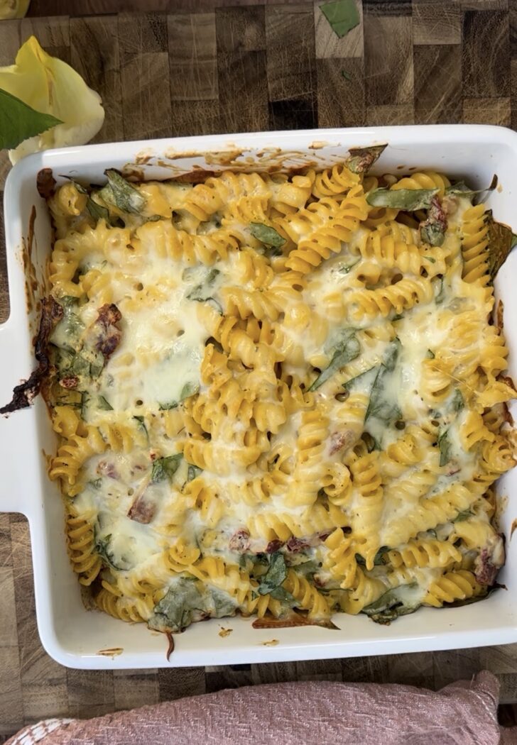 Creamy high-protein pasta bake made with rotini pasta, melted cheese, spinach, and sun-dried tomatoes baked until golden and bubbly in a white casserole dish. Cozy protein-packed dinner recipe perfect for meal prep, weeknight dinners, or healthy comfort food.