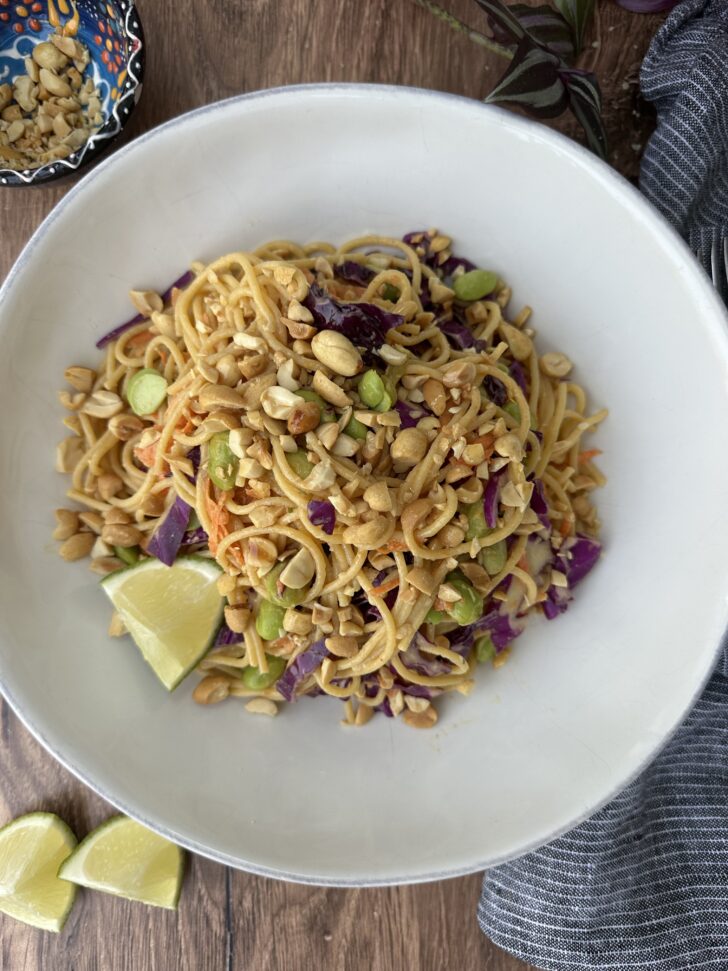 Overhead photo of a high-protein peanut noodle salad served in a white bowl with spaghetti noodles, shredded cabbage, carrots, edamame, crushed peanuts, sesame seeds, and lime wedges on the side.