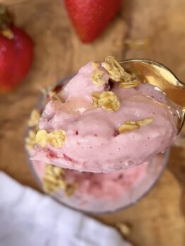 High protein strawberry frozen yogurt topped with granola and honey in a glass bowl with a gold spoon, fresh strawberries in the background on a wooden cutting board