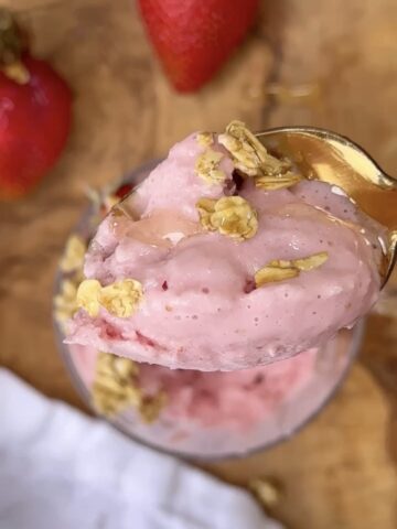 High protein strawberry frozen yogurt topped with granola and honey in a glass bowl with a gold spoon, fresh strawberries in the background on a wooden cutting board