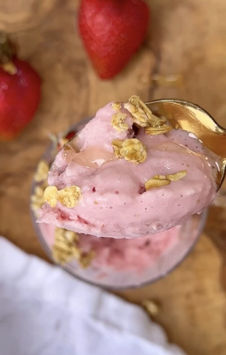High protein strawberry frozen yogurt topped with granola and honey in a glass bowl with a gold spoon, fresh strawberries in the background on a wooden cutting board