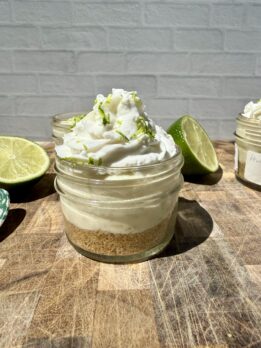 Three high-protein key lime pie cups in small glass jars on a wooden cutting board, topped with swirls of whipped cream and fresh lime zest. Freshly sliced limes sit beside the jars, creating a bright, summery dessert scene with a cozy kitchen backdrop.