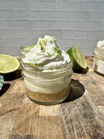 Three high-protein key lime pie cups in small glass jars on a wooden cutting board, topped with swirls of whipped cream and fresh lime zest. Freshly sliced limes sit beside the jars, creating a bright, summery dessert scene with a cozy kitchen backdrop.
