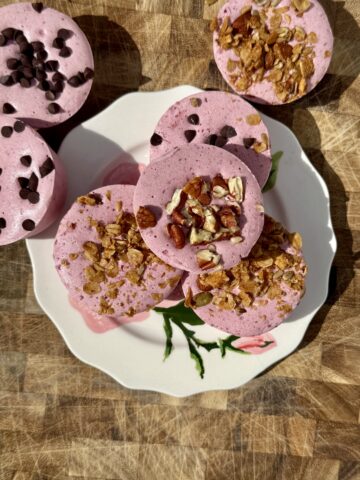 Frozen cherry yogurt cups topped with mini chocolate chips, chopped pecans, and crunchy granola arranged on a white floral plate. The pink high-protein frozen yogurt cups are photographed in natural sunlight on a rustic wooden cutting board, highlighting their creamy texture and colorful toppings. Perfect as a healthy summer snack, high-protein dessert, or post-workout treat.