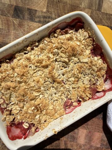 A homemade strawberry rhubarb protein crumble before baking, layered with sliced strawberries and rhubarb and topped with a rustic mixture of rolled oats, vanilla protein powder, sliced almonds, and shredded coconut in a white ceramic baking dish. The crumble sits on a wooden cutting board in bright natural sunlight, highlighting the vibrant red fruit and textured topping.