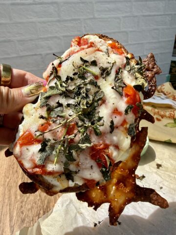 High-protein cottage cheese pizza toast made with sourdough bread, roasted cherry tomatoes, melted mozzarella cheese, and crispy baked basil. The toast is held up in natural sunlight, showing crispy cheese edges and fresh pizza-inspired toppings.