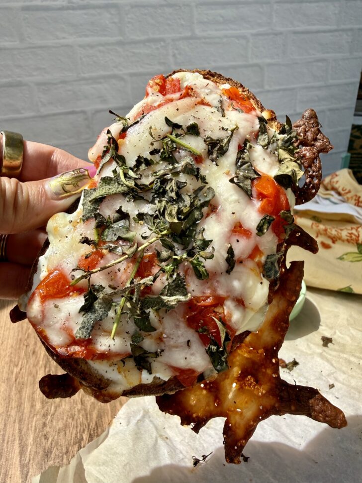 High-protein cottage cheese pizza toast made with sourdough bread, roasted cherry tomatoes, melted mozzarella cheese, and crispy baked basil. The toast is held up in natural sunlight, showing crispy cheese edges and fresh pizza-inspired toppings.
