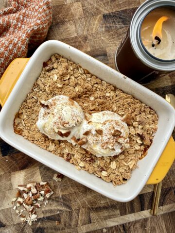 A cozy pumpkin protein crisp baked in a white ceramic baking dish with mustard yellow handles, topped with two scoops of vanilla ice cream, chopped pecans, and a dusting of cinnamon. The crisp features a golden oat crumble over a pumpkin filling and is styled on a rustic wooden cutting board beside a lit candle and a warm-toned fall kitchen towel, creating an inviting autumn-inspired dessert scene.