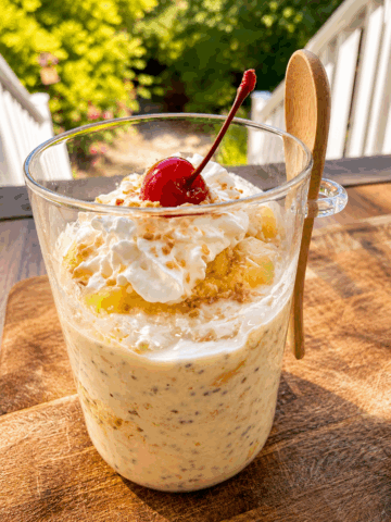 High-protein pineapple upside-down cake overnight oats layered with creamy vanilla oats, juicy pineapple, whipped topping, toasted coconut, and a maraschino cherry, served in a clear glass mug on a wooden table outdoors.