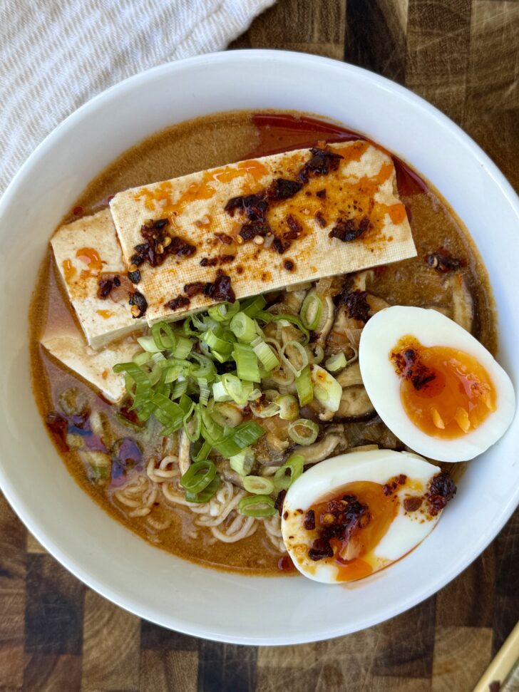 Overhead view of a bowl of creamy high-protein miso ramen with a rich golden-brown broth, curly ramen noodles, sliced shiitake mushrooms, two seared tofu planks, a jammy soft-boiled egg cut in half with a vibrant orange yolk, chopped green onions, and chili crisp drizzled over the top. The ramen is served in a white bowl on a wooden cutting board, creating a cozy, restaurant-style presentation.