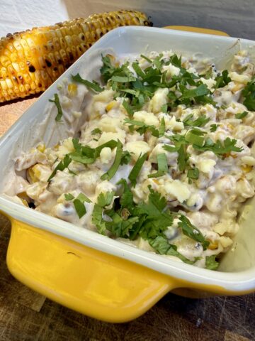 Close-up of a creamy high-protein street corn dip made with blended cottage cheese and Greek yogurt, mixed with charred grilled corn, and topped with fresh cilantro and crumbled cotija cheese. Two grilled ears of corn with smoky char marks sit in the background, highlighting this healthy summer appetizer served in a bright yellow baking dish.