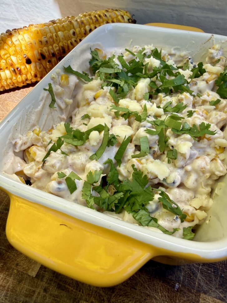 Close-up of a creamy high-protein street corn dip made with blended cottage cheese and Greek yogurt, mixed with charred grilled corn, and topped with fresh cilantro and crumbled cotija cheese. Two grilled ears of corn with smoky char marks sit in the background, highlighting this healthy summer appetizer served in a bright yellow baking dish.
