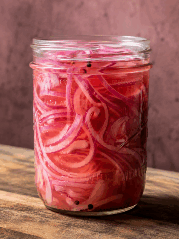 Homemade quick pickled red onions in a glass mason jar with apple cider vinegar brine and black peppercorns on a rustic wooden surface.