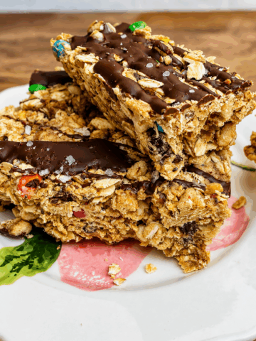 Close-up of thick no-bake protein monster cookie bars made with rolled oats, peanut butter, mini chocolate chips, colorful mini M&M's, and a dark chocolate drizzle. The chewy high-protein bars are stacked on a white plate with flaky sea salt on top, making an easy meal prep snack or healthy dessert.