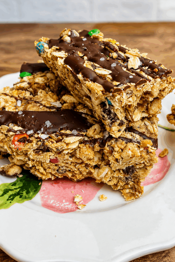 Close-up of thick no-bake protein monster cookie bars made with rolled oats, peanut butter, mini chocolate chips, colorful mini M&M's, and a dark chocolate drizzle. The chewy high-protein bars are stacked on a white plate with flaky sea salt on top, making an easy meal prep snack or healthy dessert.
